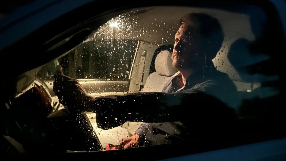 David Sparrow seated in a car at night behind a rain-covered window