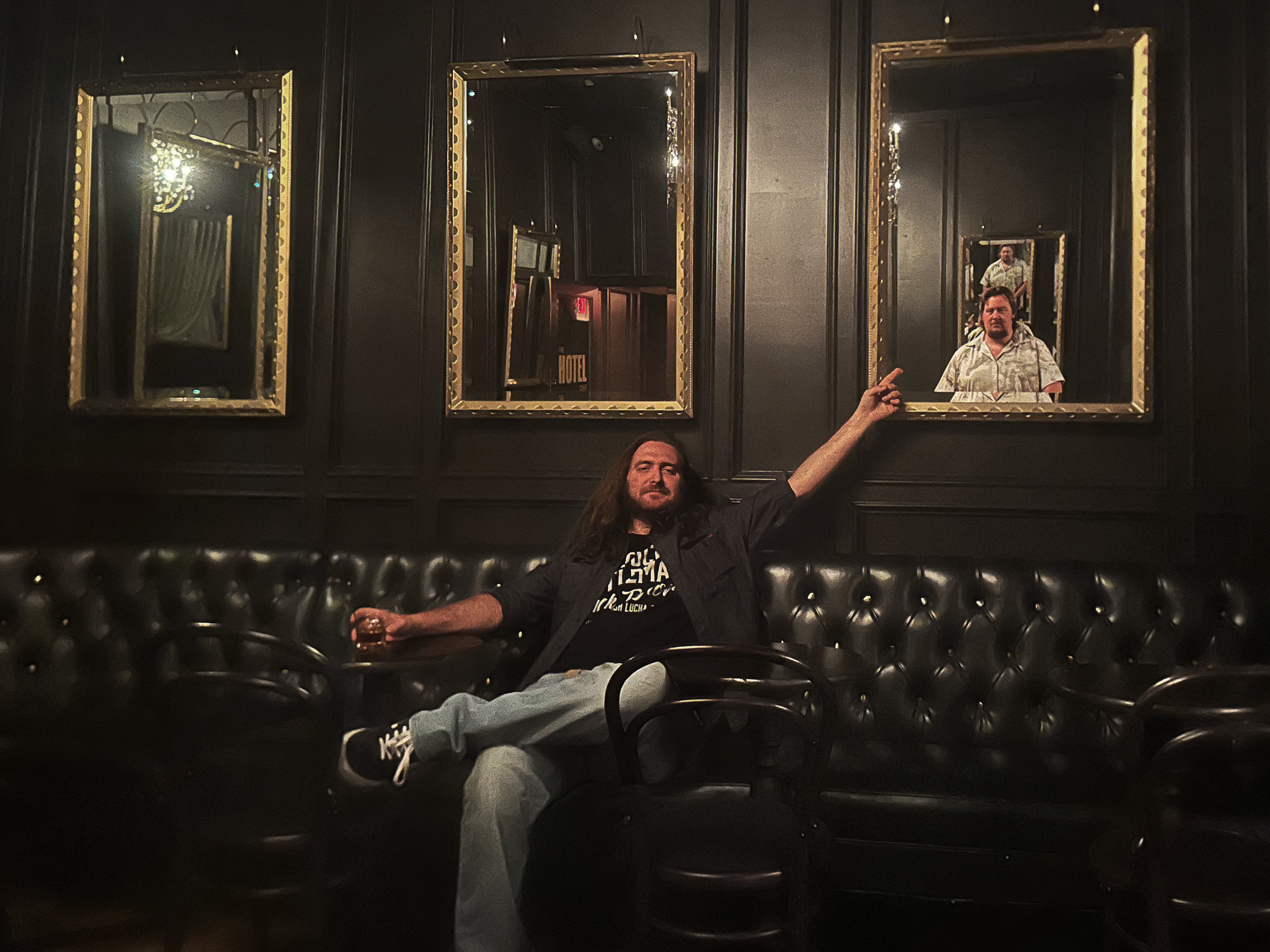 A man seated on a dark leather sofa beneath gold-framed mirrors, with another man reflected behind him
